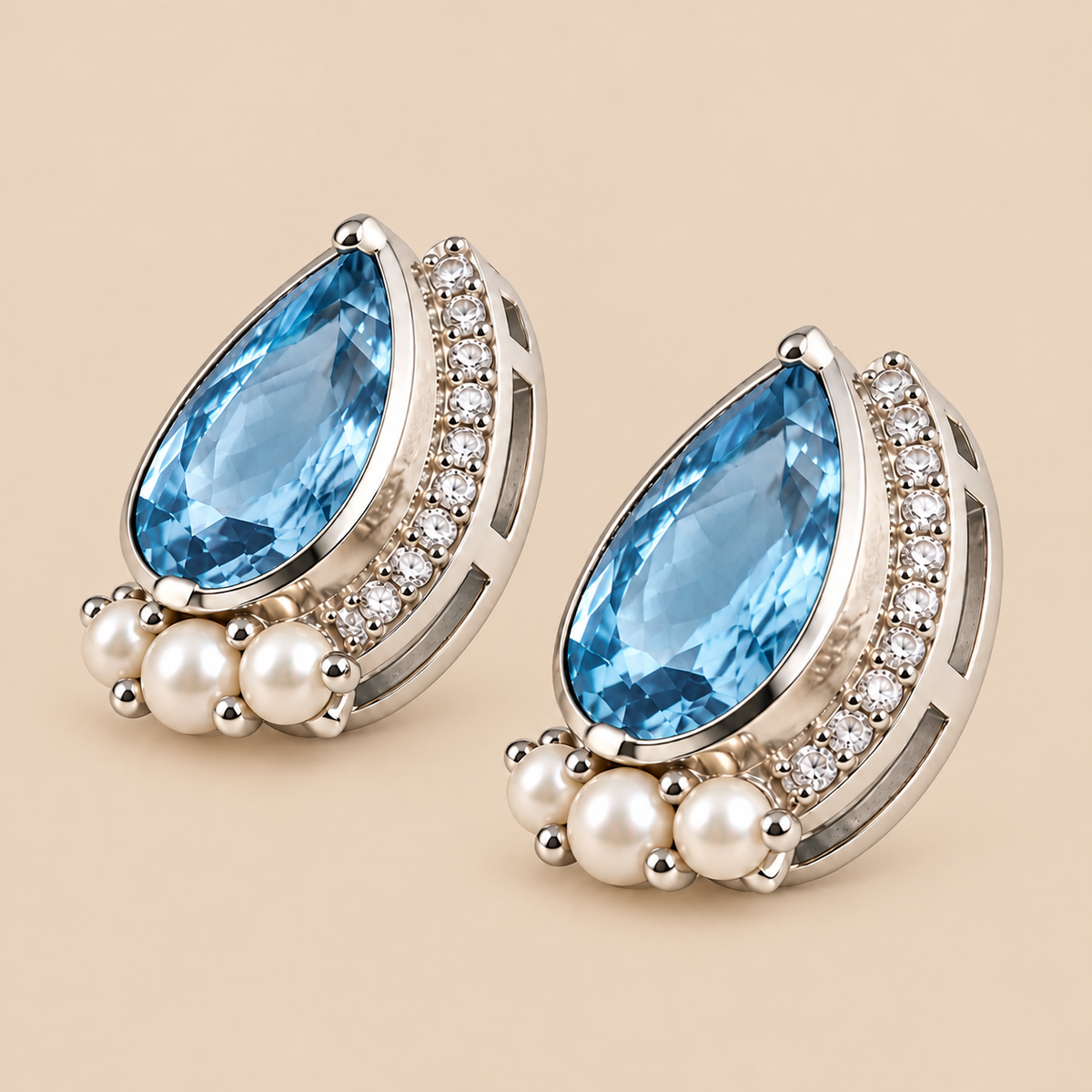 A pair of teardrop-shaped earrings featuring swiss blue topaz gemstones, surrounded by clear pavé stones and accented with small white pearls, set in a silver-toned metal on a warm beige background.