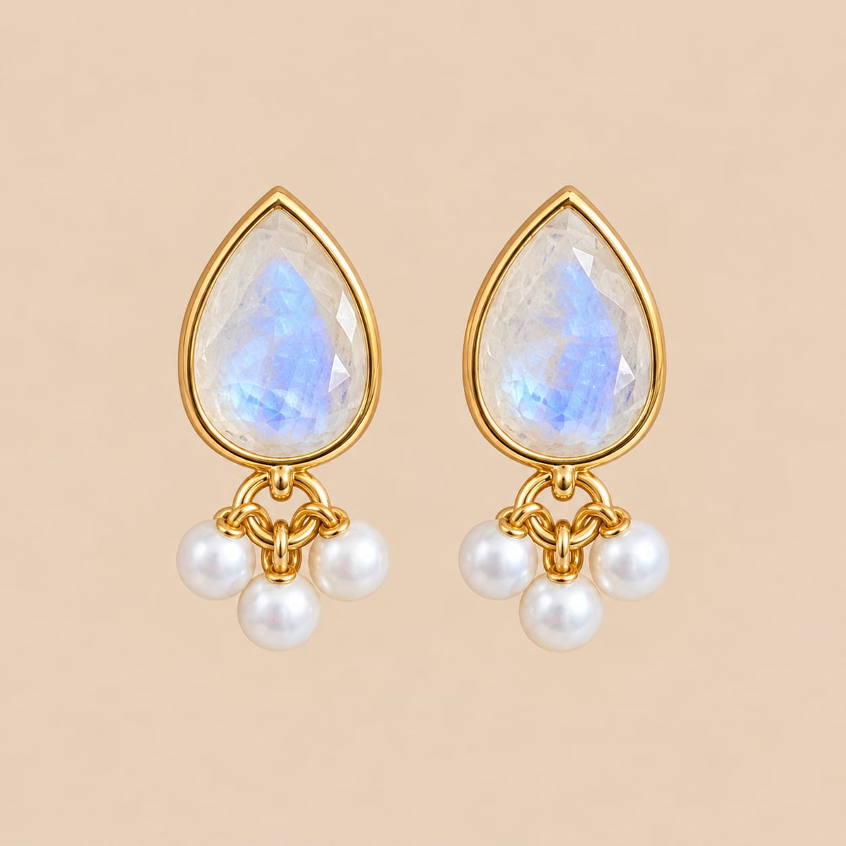 A pair of gold teardrop-shaped dangle earrings featuring iridescent white moonstone tops and three small white pearl drops below, set against a solid beige background.