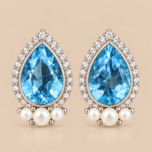 A pair of teardrop-shaped earrings featuring swiss blue topaz gemstones, surrounded by clear pavé stones and accented with small white pearls, set in a silver-toned metal on a warm beige background.